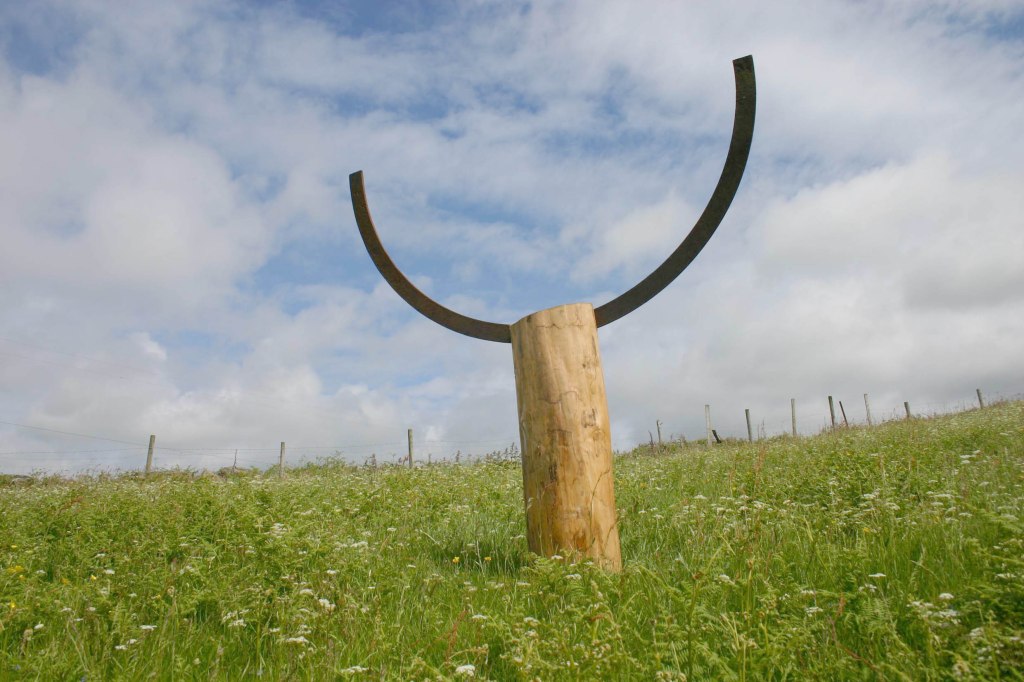 Sculpture made of a tall tree trunk wood base with an ark of cast iron slotted into the top reaching upwards. It stands in a field of long grass and wild flowers.
