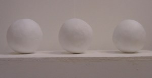 Three white, spherical clay sculptures in a line on a white plinth