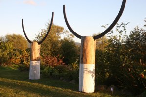 Two sculptures, tree trunks, half painted white with cast iron semi circles arching upwards from the top. In a garden with trees and shrubs.